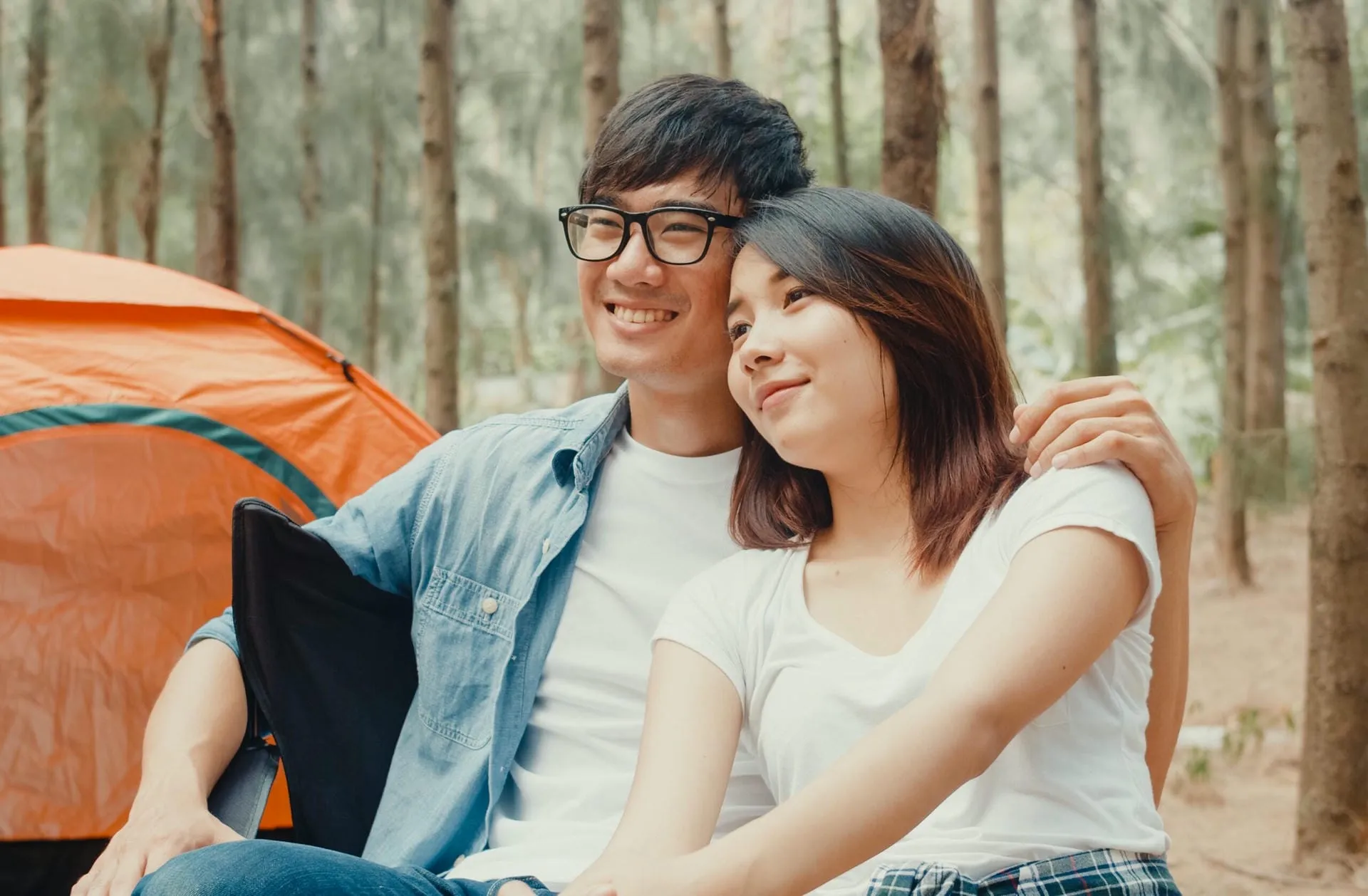 Young asia campers couple sitting in chairs by tent in forest. Male and female traveler relaxing and talking on a summer day at campsite. Outdoor activity, adventure travel, or holiday vacation.